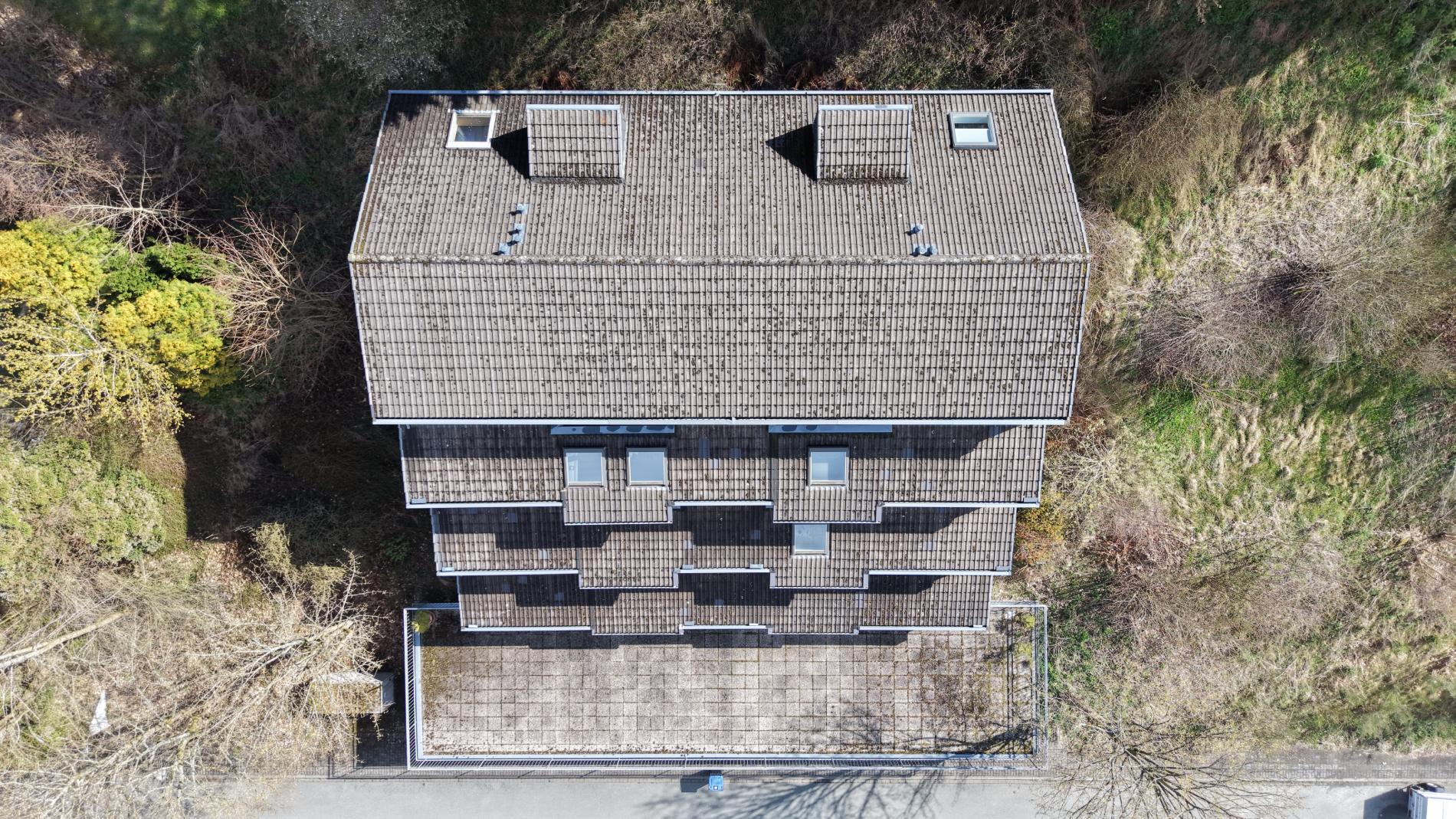 Drohnen-Übersichtsaufnahme eines Mehrfamilienhauses aus der Vogelperspektive. Sichtbar sind die gesamte Dachfläche mit Ziegeldeckung, zwei Dachfenster, zwei Gauben, Schornsteinaufbauten und die Dachterrasse im Vordergrund.
