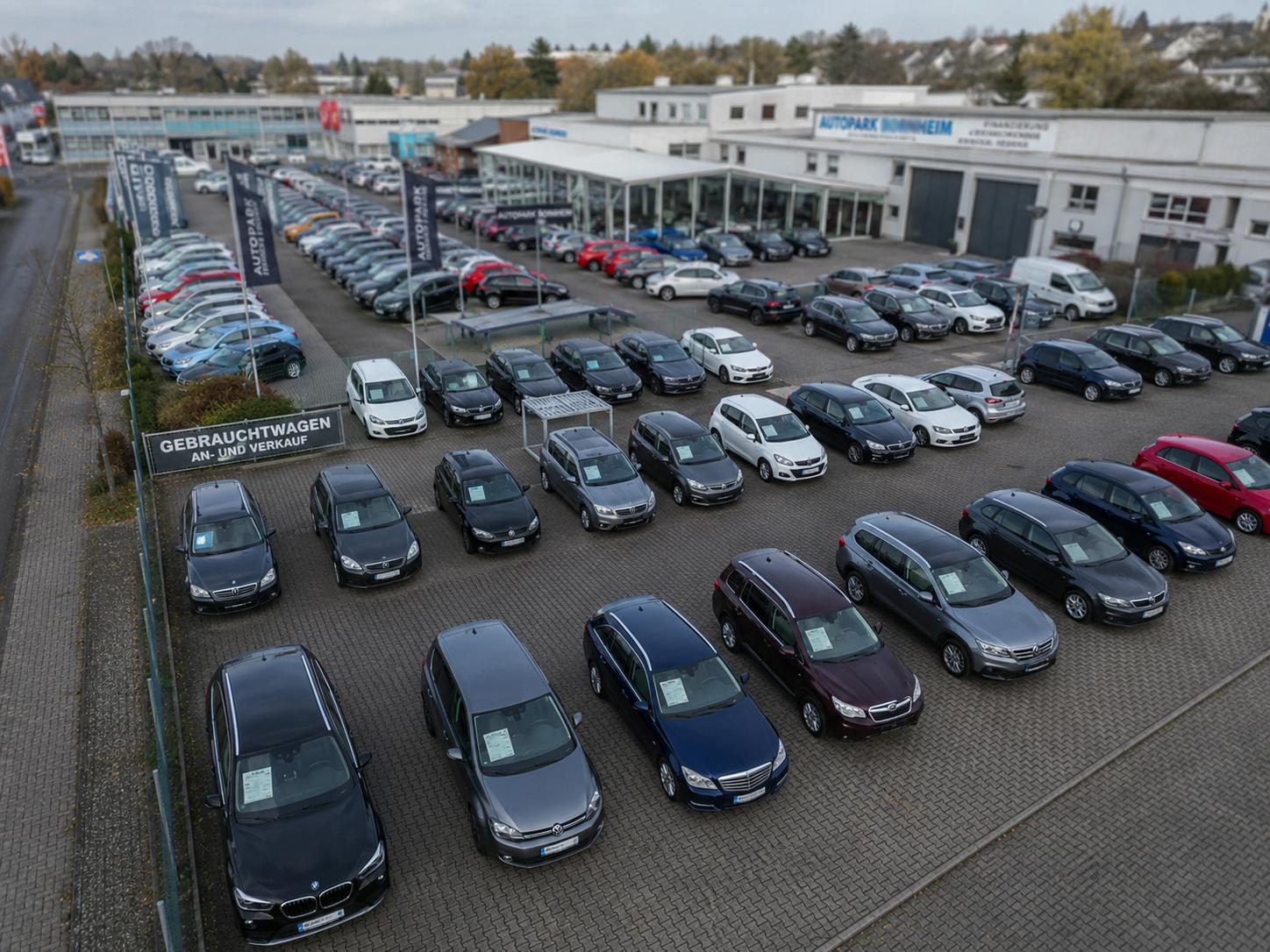Drohnen-Übersichtsaufnahme eines Gebrauchtwagen-Handels. Aus der Vogelperspektive sind etwa 80 geordnet aufgestellte Fahrzeuge sichtbar, das Verkaufsgebäude mit großer Glasfront und Fahnenmasten von 'Autopark Bornheim'. Im Hintergrund weitere Gewerbegebäude und Vegetation.