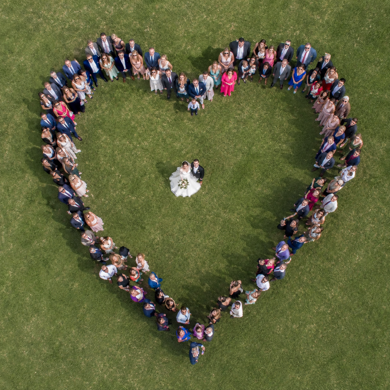 Drohnen-Aufnahme aus der Vogelperspektive: Hochzeitspaar steht im Zentrum einer Wiese, die Braut in weißem Brautkleid mit Brautstrauß, der Bräutigam im dunklen Anzug. Um das Paar bilden alle Hochzeitsgäste eine perfekte Herzform – mehrere Dutzend festlich gekleidete Personen, die nach oben in die Kamera blicken.