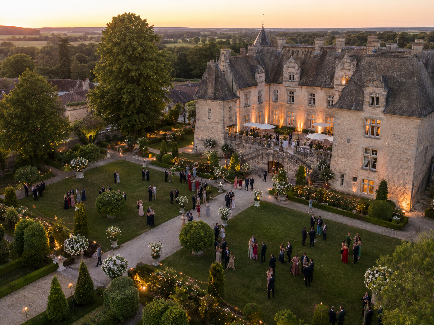 Drohnen-Übersichtsaufnahme einer Hochzeitsgesellschaft im Schlosspark zur goldenen Abendstunde. Im Zentrum ein historisches Schloss mit Sandsteinfassade, hellen Turmspitzen und angestrahlten Fenstern. Davor ein formaler Garten mit symmetrischen Wegen, geschnittenen Buchsbaumkugeln, Blumenarrangements und Lichtgirlanden, in dem etwa 60 elegant gekleidete Gäste verteilt stehen. Im Hintergrund weite Felder und Wälder im warmen Sonnenuntergangslicht.